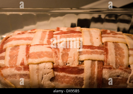 Polpettone crudo avvolto con il bacon in una cesta modello di armatura seduto in un chiaro in Pyrex teglia su una cucina piano cottura Foto Stock