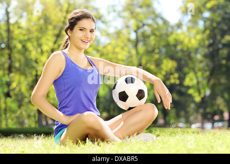 Giovane atleta donna seduta su un prato verde e trattenimento di un pallone da calcio in un parco Foto Stock