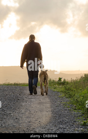 Una giovane donna a piedi un cane su un percorso pietroso, allontanandosi dalla telecamera nell'ambiente SUN. Foto Stock