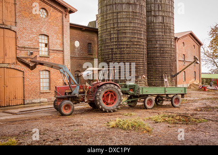 Il trattore e il rimorchio parcheggiato in un cortile di fronte ad una grande stalla multipiano e silos cilindrici con un famer appena esce il trattore dopo provenienti dai campi Foto Stock