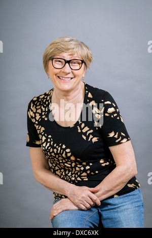 Attraente bionda senior donna che indossa gli occhiali seduto con mani giunte in una posizione rilassata sorridente alla fotocamera, su grigio Foto Stock