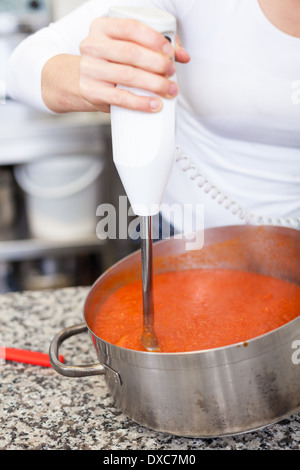 Lo chef donna sbattere bollito di pomodoro in una pentola per una loro riduzione in una purea di base per zuppe o salse con un dispositivo azionato a batteria w Foto Stock