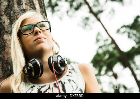 Ritratto di una ragazza che indossa occhiali, in piedi accanto ad albero nel parco, con le cuffie intorno al collo, guardando verso l'alto, Germania Foto Stock
