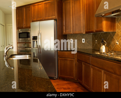 Primo piano di una passerella dietro Isola per cucina con elettrodomestici in acciaio inox, fornello a gas a superfici in pietra e legno di ciliegio Foto Stock