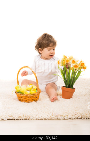 Il Toddler ragazzo con cesto di Pasqua con le uova di organizzare i narcisi in una pentola e seduti su un tappeto Foto Stock