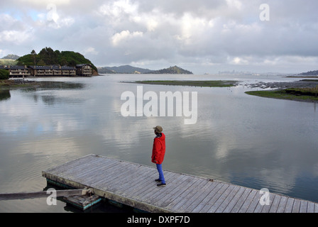 Woman in Red coat stand su dock guardando la riflessione di nuvole sulla Baia di San Francisco Foto Stock