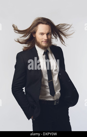 Un uomo barbuto, modello maschile, capelli lunghi in posa di un abito nero e cravatta guardando in giù con il movimento dei capelli, uno degli uomini del concetto di moda Foto Stock