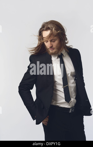 Un uomo barbuto, modello maschile, capelli lunghi in posa di un abito nero e cravatta guardando in giù con il movimento dei capelli, uno degli uomini del concetto di moda Foto Stock