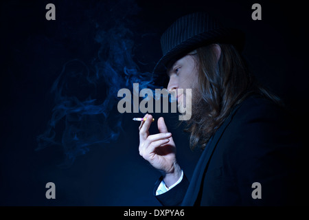 Un barbuto lunghi capelli uomo che indossa un cappello Fedora e tuta fumatori, un profilo laterale verticale con il sentiero di fumo, di un concetto creativo Foto Stock