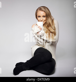 Carino e bella giovane donna riscaldino con una calda tazza di tè o caffè. Girato su sfondo grigio. Modello bionda. Foto Stock