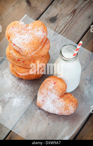 Una pila di zucchero a velo cosparso, ciliegia a forma di cuore ciambelle, con una bottiglia di latte pieno di latte e un vecchio carta paglia. Foto Stock