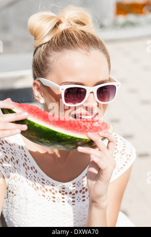 Ragazza bionda con il filtro bow tie capelli in bianco abiti estivi indossando occhiali da sole morde succosa anguria Foto Stock