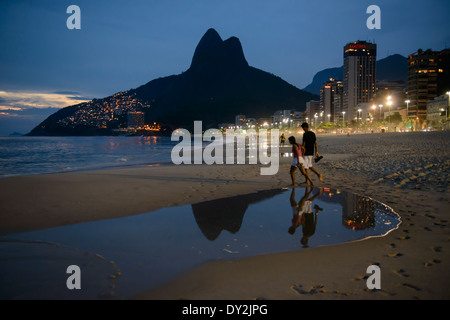 Giovane a piedi lungo la spiaggia di Ipanema di notte,con picchi gemelli di due fratelli di montagna a Leblon, in background, Rio de Janeiro, B Foto Stock