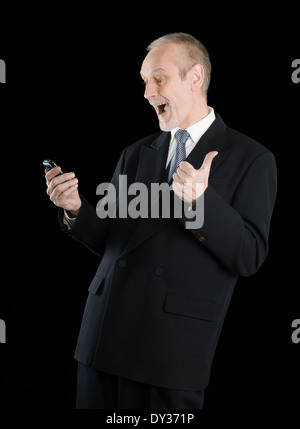 Happy businessman indossando un vestito nero, sorridente e la lettura di sms sul cellulare, con pollice in alto, su sfondo nero Foto Stock