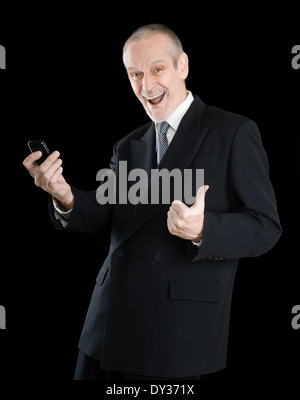 Happy businessman indossando un vestito nero, sorridente e la lettura di sms sul cellulare, con pollice in alto, su sfondo nero Foto Stock