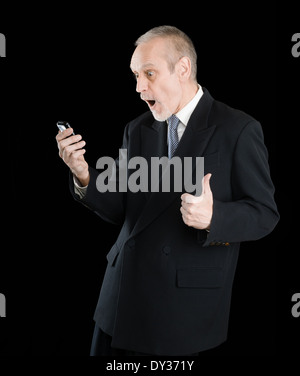 Happy businessman indossando un vestito nero, sorridente e la lettura di sms sul cellulare, con pollice in alto, su sfondo nero Foto Stock