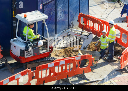 Vista aerea tre squadre di uomini che indossano elmetto e giacca ad alta visibilità utilizzando miniescavatore scavatore scavare su pavimentazione dietro barriere rosse Londra UK Foto Stock