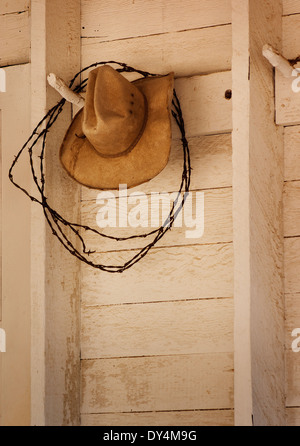 Un vecchio cappello da cowboy appesi a un picchetto in legno con filo spinato su un bianco al di fuori del muro del granaio Foto Stock