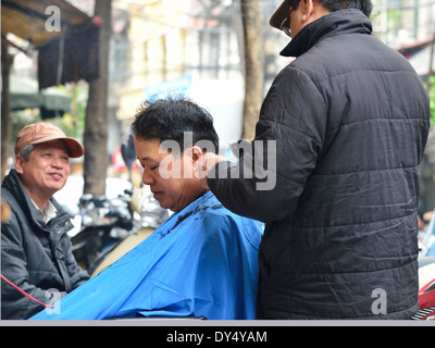 Barbiere di strada sul marciapiede con il cliente città vecchia Hanoi, Vietnam la rasatura dei capelli con azionato a batteria clippers Foto Stock