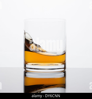 Rocce di un vetro con il Whisky si siede in cima ad una superficie riflettente su uno sfondo bianco; un'onda in vetro mostra il movimento Foto Stock