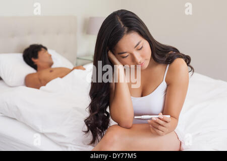 Donna seduta sul letto azienda test di gravidanza come fidanzato dorme Foto Stock