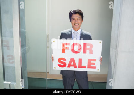 Fiducioso agente immobiliare in piedi davanti alla porta di casa che mostra per la vendita segno Foto Stock
