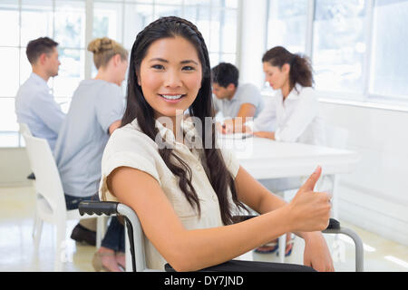 Imprenditrice casual in carrozzella sorridente in telecamera con il team dietro di lei Foto Stock