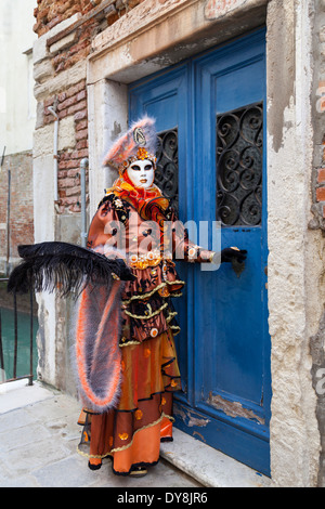 Costume di Carnevale di Venezia, scatto completo di donna mascherata in arancio abito storico fantasia apertura porta blu durante il Carneivale di Venezia, Italia Foto Stock