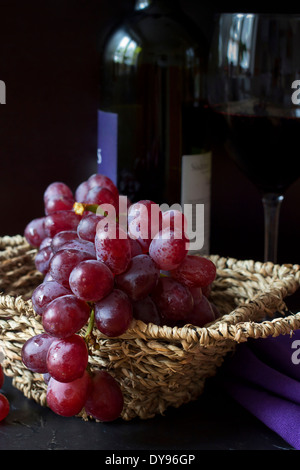 Cesto in Vimini di uve rosse (Vitis vinifera), vino rosso in vetro e bottiglia in background Foto Stock