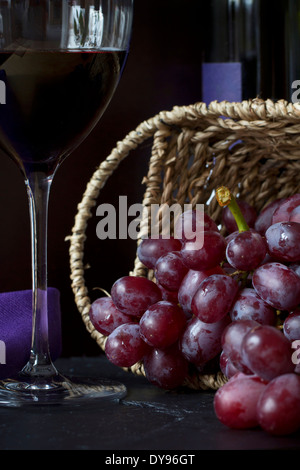 Cesto in Vimini di uve rosse (Vitis vinifera), vino rosso in vetro e bottiglia in background Foto Stock
