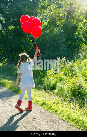 Bambina con un mucchio di palloncini rossi a camminare su una strada di campagna in un verde paesaggio estivo con nebuloso editing vintage Foto Stock