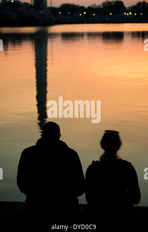 WASHINGTON DC - i fiori di ciliegio sono visti in lontananza attraverso il bacino delle maree mentre il riflesso del monumento a Washington appare nell'acqua all'alba. Gli alberi in fiore sono al centro dell'annuale National Cherry Blossom Festival, che celebra il dono 1912 degli alberi provenienti dal Giappone. Due persone vengono sagomate in primo piano mentre guardano l'alba. Foto Stock