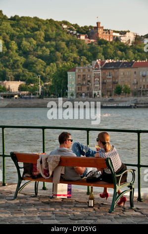 Un giovane si siede su una panchina e gode di un buon bicchiere di vino mentre guardando il Fiume Danubio al tramonto a Budapest, Ungheria Foto Stock