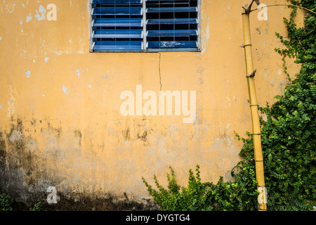 La facciata della casa di età. Parete di grungy e incrinato stucco giallo blu con finestra ritagliata. Vecchie rotture di bambù. Foto Stock