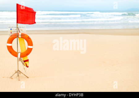 Vita dispositivi di salvataggio appesa sul rack in legno con bandiera rossa e la tavola di avvertimento 'no life-guardia' sulla spiaggia vuota. Foto Stock