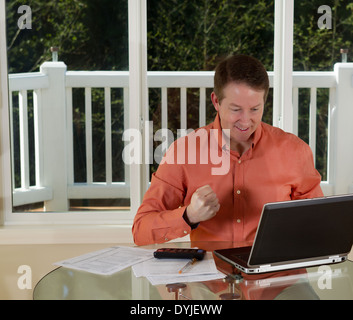 Foto di uomo maturo che mostra la felicità mentre si lavora da casa con il computer portatile, calcolatrice e carte sulla parte superiore della tavola Foto Stock