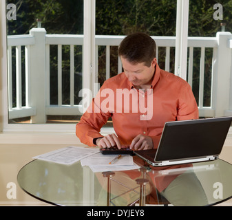 Foto di uomo maturo utilizzando la calcolatrice, mentre lavora a casa con il computer portatile, e documenti sulla parte superiore della tavola e grandi finestre Foto Stock