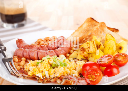 La deliziosa prima colazione inglese. Fagioli, salsicce e uova strapazzate su piastra bianca con la forcella. Caffè in background. Foto Stock