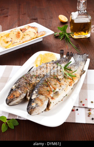 Due trote alla griglia sulla piastra bianca con pezzi di limone, patate e olio d'oliva sul panno da cucina sul tavolo di legno. Foto Stock