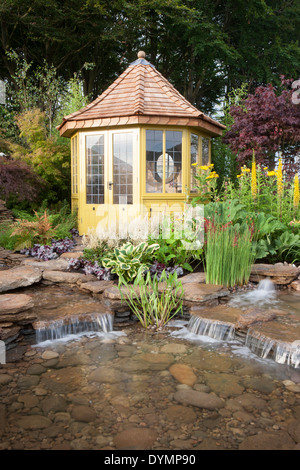 Un giardino suburbano di paese in estate con una casa estiva cabina piccolo stagno acqua caratteristica con cascata e fiori misti colorati confine UK Foto Stock