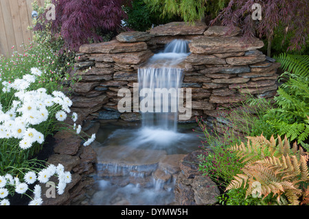 il giardino con una cascata d'acqua presenta piccole cascate rocciose con pareti in pietra asciutta in un giardino che ospita alberi acer acri felci Regno Unito Foto Stock