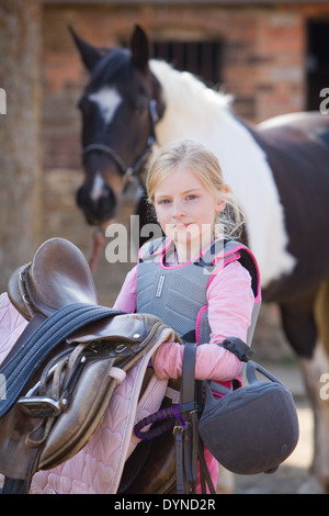 Una giovane ragazza in piedi con un cavallo al di fuori in un cantiere stabile in una giornata di sole e guardando la telecamera Foto Stock