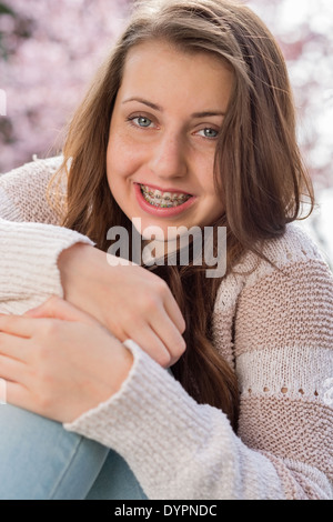 Sorridente ragazza adolescente con bretelle abbracciando le ginocchia all'aperto Foto Stock