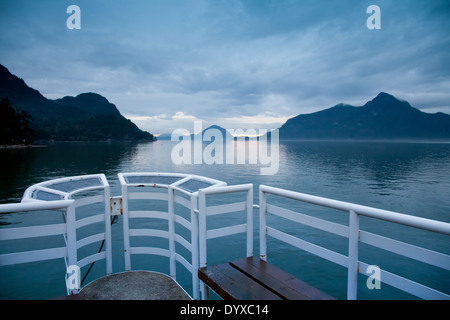 Visualizzare attraverso la calma acqua in ingresso del metallo bianco pier e il gantry con circondante Misty Mountains, drammatica sky Foto Stock