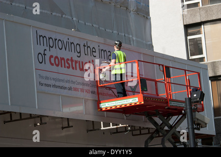 Costruttore che erige un cartello "Improving the Image of Construction" per lo schema dei costruttori premurosi utilizzando una piattaforma di lavoro aerea con elevatore a forbice (ciliegia picchettatrice) a Londra, Regno Unito. Promuovere le migliori pratiche e la sicurezza nei progetti di costruzione urbana. Foto Stock