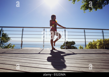 Ritratto di giovane modello femminile in bikini rosso in piedi da una ringhiera con un bicchiere di cocktail che guarda lontano. Giovane donna in balcone Foto Stock