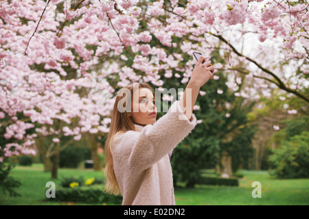 Giovane donna caucasica dipinge sbocciare fiori con il suo telefono cellulare. Donna attraente fotografare fiori presso il Parco di primavera Foto Stock