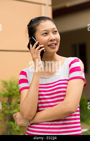 Una bella giovane donna asiatica a parlare su un telefono intelligente e sorridente cercando, in piedi all'esterno. Foto Stock