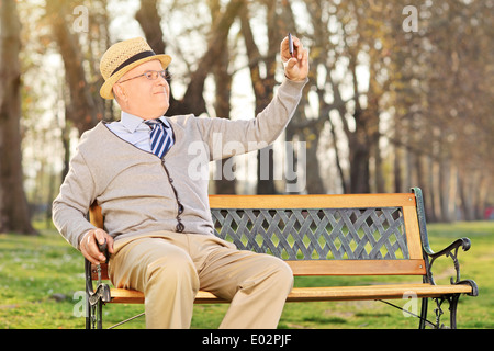 Anziano gentiluomo prendendo un fumetto selfie cellulare, seduto su un banco di lavoro in posizione di parcheggio Foto Stock