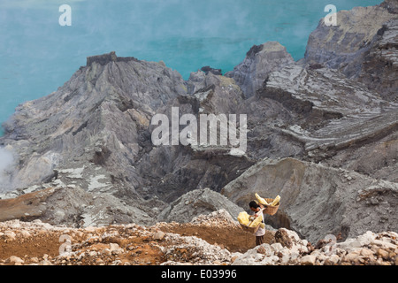 Uomo che porta cestelli laden con blocchi di zolfo. Acido turchese del lago del cratere è visibile al di sotto, Kawah Ijen, East Java, Indonesia Foto Stock